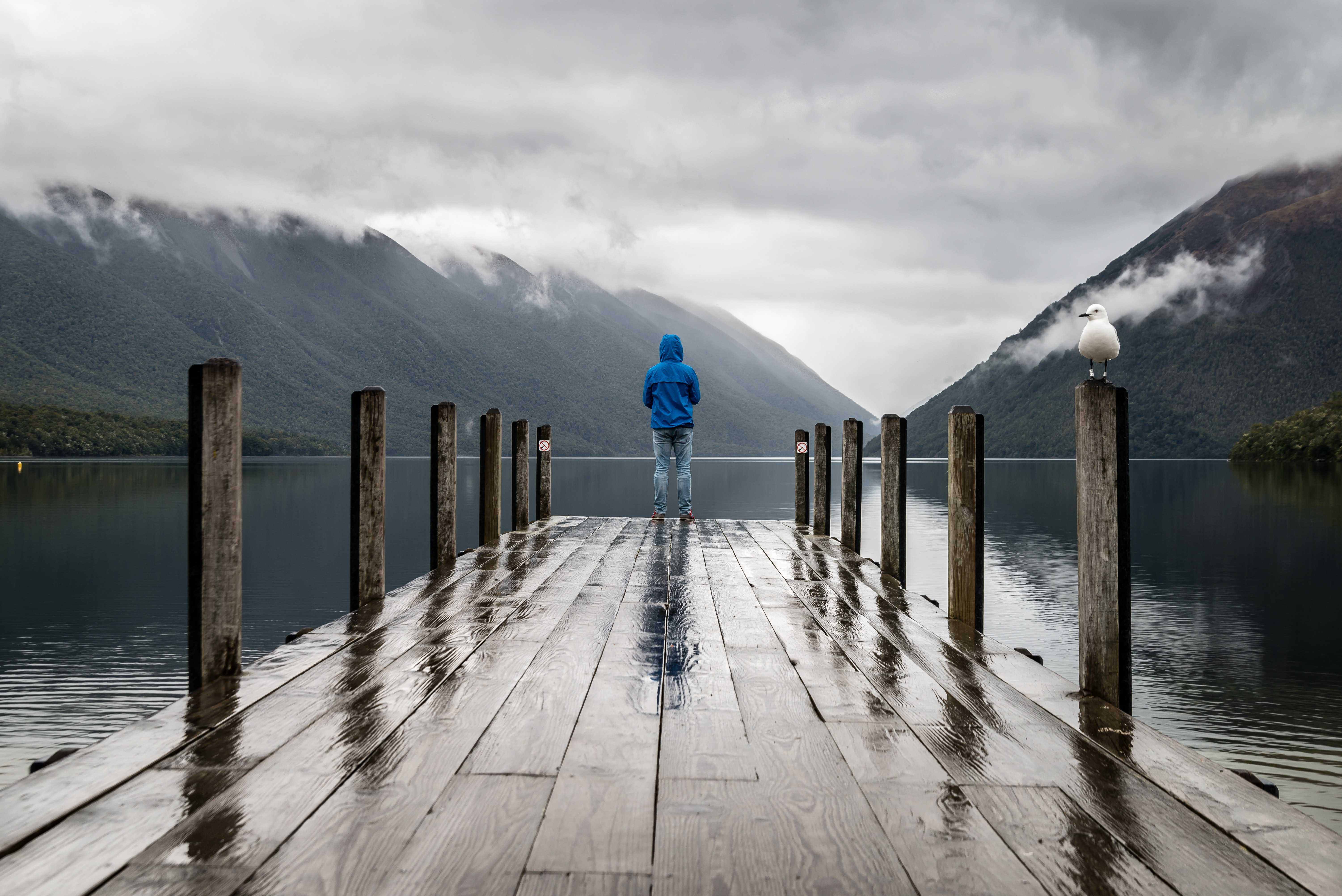Boy standing at end of wet pier overlooking still lake with mountains in background
